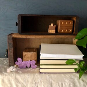 Puede incluir: Un cajón de madera con una pila de libros encima. El cajón está abierto y hay bloques de madera y una pequeña casa de madera en su interior. La palabra "LOVE" está escrita en letras de fieltro morado en la parte delantera del cajón.