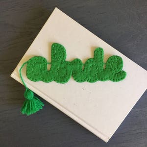 Puede incluir: Marca páginas de fieltro verde con la palabra "bride" bordada en hilo blanco. El marcapáginas tiene un borla verde.