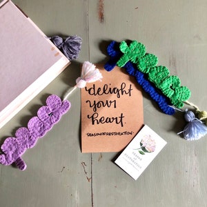 Puede incluir: Un conjunto de cuatro marcapáginas de tela coloridos con borlas. Los marcapáginas tienen forma de corazón y tienen un diseño cosido. Los marcapáginas están dispuestos en una tarjeta de papel marrón con el texto "delight your heart.  SEASON OF RESTORATION."