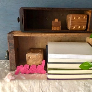 Puede incluir: Un mueble de cajones de madera con dos estantes. El estante superior tiene una casa de madera y un dado de madera. El estante inferior tiene una pila de libros con un marcapáginas de fieltro rosa que dice "leer".