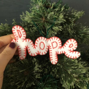 Puede incluir: Adorno navideño de fieltro blanco con la palabra "HOPE" bordada en hilo rojo.