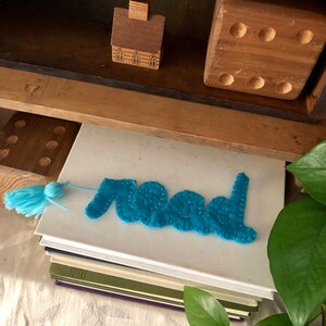 Puede incluir: Un marcapáginas de fieltro azul con la palabra "leer" bordada en hilo blanco. El marcapáginas está encima de una pila de libros.