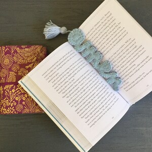 Puede incluir: Un marcapáginas de fieltro azul claro con un borla. El marcapáginas tiene forma de una serie de bucles conectados. El marcapáginas está sobre un libro abierto con texto sobre el arrepentimiento y la fe.
