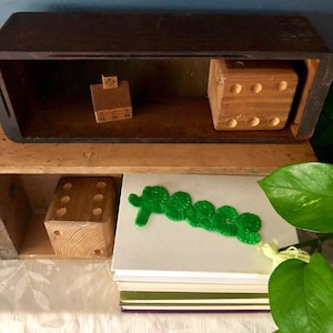 Puede incluir: Una caja de madera con dos estantes. El estante superior tiene una pequeña casa de madera y un bloque de madera con agujeros. El estante inferior tiene un bloque de madera con agujeros y la palabra "paz" en fieltro verde.