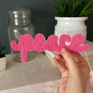 Puede incluir: Una palabra de fieltro rosa "peace" con costuras blancas se sostiene frente a un tarro de cristal transparente y una maceta blanca con una planta verde. La palabra está en cursiva. Fondo con mantel blanco.