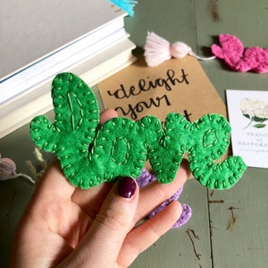 Puede incluir: Letras de fieltro verde "LOVE" con costuras blancas, sostenidas en una mano sobre un fondo de madera verde. Las letras forman parte de un conjunto de marcapáginas de fieltro con la frase "delight your heart" escrita en un fondo de papel marrón.