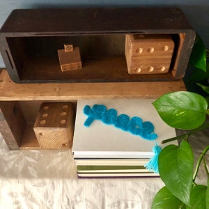 Puede incluir: Una caja de madera con dos estantes. El estante superior tiene una pequeña casa de madera y un bloque de madera con seis lados. El estante inferior tiene un bloque de madera con seis lados y un marcapáginas de fieltro azul en forma de signo de paz. El marcapáginas tiene un borla azul.