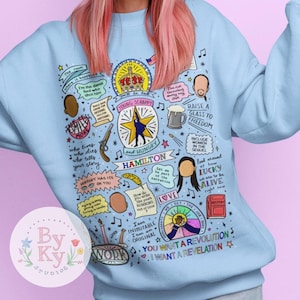 Peut inclure: Sweat-shirt bleu clair avec un imprimé graphique coloré représentant des paroles et des personnages de la comédie musicale Hamilton. Le graphique comprend les mots "Young, Scrappy, and Hungry", "Hamilton", "Raise a Glass to Freedom", "You Want a Revolution, I Want a Revelation", et "I am inimitable, I am an original".