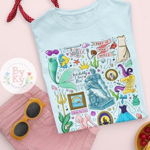 Pu&ograve; includere: Maglietta blu chiaro con un'illustrazione colorata di vari personaggi e oggetti del film La Sirenetta. L'illustrazione include Ariel, il principe Eric, Sebastian, Ursula e altri elementi iconici. Il testo "Part of Your World" &egrave; incluso nel design.