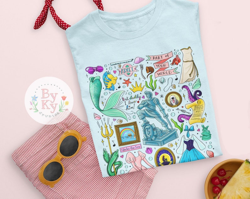 Pu&ograve; includere: Maglietta blu chiaro con un'illustrazione colorata di vari personaggi e oggetti del film La Sirenetta. L'illustrazione include Ariel, il principe Eric, Sebastian, Ursula e altri elementi iconici. Il testo "Part of Your World" &egrave; incluso nel design.