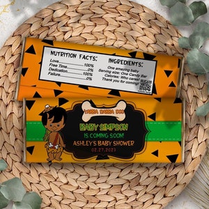 Puede incluir: Un diseño de envoltorio de barra de caramelo imprimible para un baby shower. El envoltorio es naranja y negro con una imagen de dibujos animados de un bebé con un disfraz de cavernícola. El texto en el envoltorio dice "Wabba Dabba Doo Baby Simpson is Coming Soon! Ashley's Baby Shower 02.27.2023".