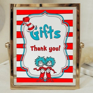 May include: A framed sign with a red and white striped background and the word "Gifts" in turquoise. The sign also includes the text "Thank you!" and illustrations of characters. The frame is gold and the sign is 20.3 x 25.4 cm.