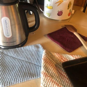 May include: A stainless steel Hamilton Beach electric kettle and a white slow cooker with vegetable illustrations sit on a countertop. Two knitted washcloths, one blue and one multi-colored, and a maroon pot holder are also visible.