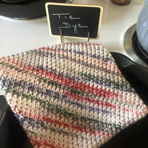 May include: A square, hand-crocheted cotton dishcloth featuring a variegated tie-dye pattern with diagonal stripes of red, blue, green, and white. A small black rectangular chalkboard sign with the handwritten white text Tie Dye stands behind it.