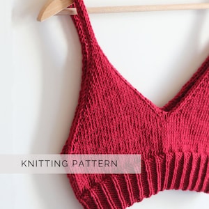 Può includere: Un top corto di maglia rosso con scollo a V e sottili spalline. Il top è appeso a un appendiabiti di legno. "Knitting Pattern" è scritto in bianco nella parte inferiore dell'immagine.