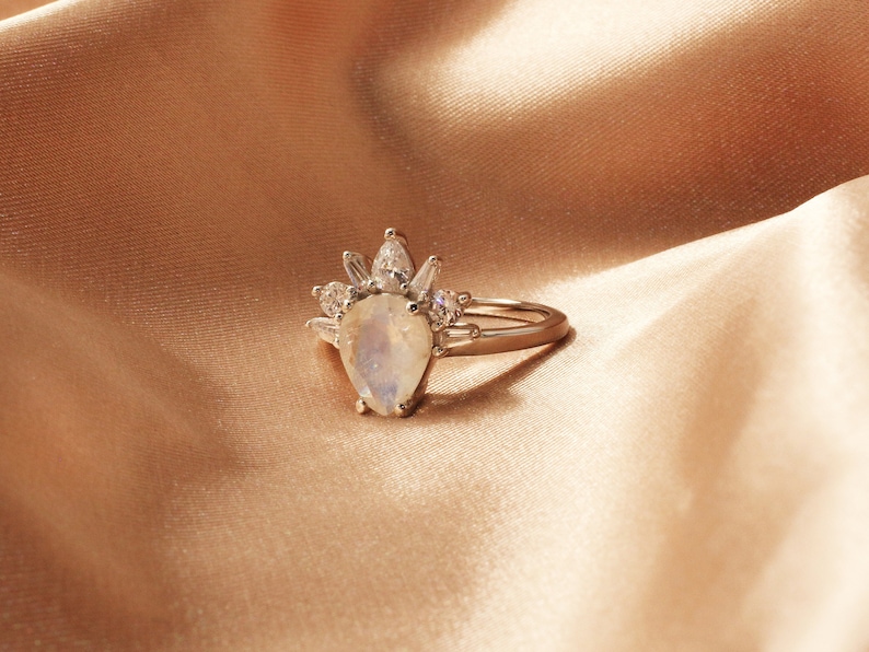 Puede incluir: Un anillo de oro con una piedra de luna en forma de pera y un halo de diamantes m&aacute;s peque&ntilde;os. El anillo est&aacute; sobre una tela suave de color melocot&oacute;n.