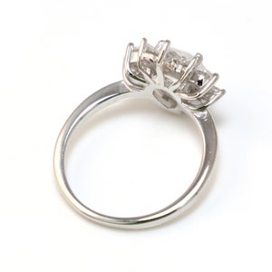 Puede incluir: Un anillo de plata con una piedra preciosa grande y transparente rodeada de piedras preciosas m&aacute;s peque&ntilde;as. El anillo est&aacute; sobre un fondo blanco.