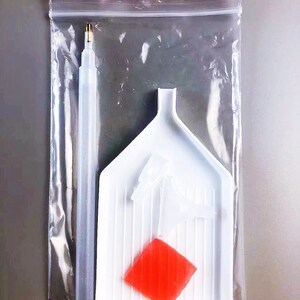 May include: A clear plastic bag containing diamond painting supplies. The supplies include a white tray with a red diamond-shaped piece, a clear plastic pen with a gold tip, and two white plastic tools.