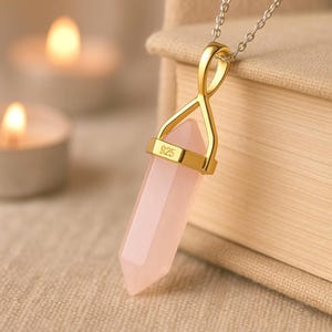 May include: A rose quartz crystal pendant necklace. The pendant is a pale pink, elongated crystal with a gold-colored metal cap marked "925". The necklace chain is silver-colored. The background includes soft lighting and a book.