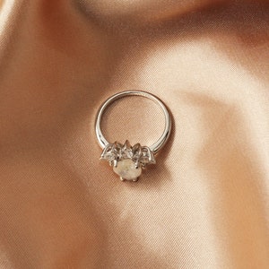Puede incluir: Un anillo de plata con una gran piedra preciosa ovalada rodeada de piedras preciosas m&aacute;s peque&ntilde;as y transparentes. El anillo est&aacute; colocado sobre una tela suave de color melocot&oacute;n.