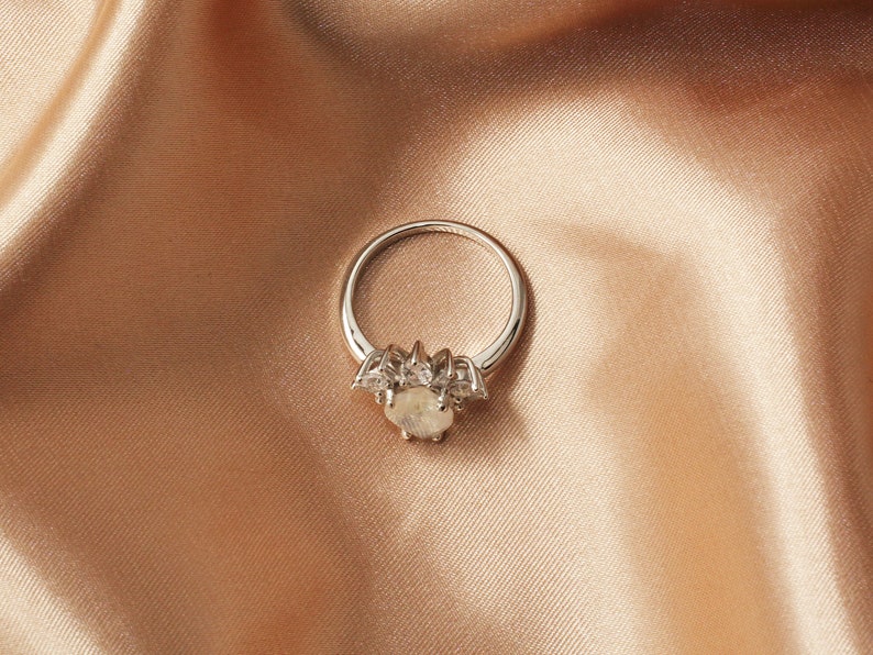Puede incluir: Un anillo de plata con una gran piedra preciosa ovalada rodeada de piedras preciosas m&aacute;s peque&ntilde;as y transparentes. El anillo est&aacute; colocado sobre una tela suave de color melocot&oacute;n.