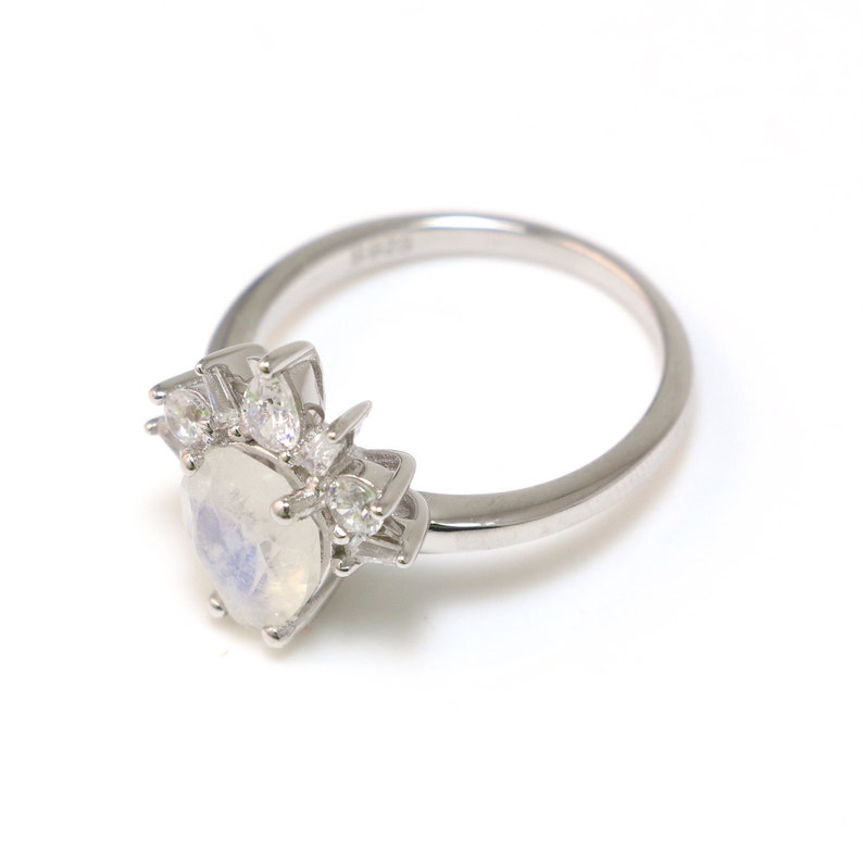 Puede incluir: Un anillo de plata con una gran piedra de luna ovalada y piedras m&aacute;s peque&ntilde;as transparentes engastadas en un dise&ntilde;o de corona.