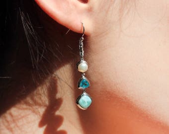 May include: A silver earring with three decorative elements: a white pearl, a blue triangular stone, and a light blue square stone. The earring is hanging near a person's ear.