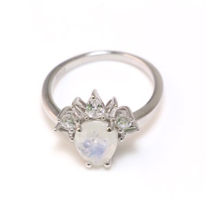 Puede incluir: Un anillo de plata con una piedra central de piedra lunar nacarada y piedras m&aacute;s peque&ntilde;as y claras alrededor.