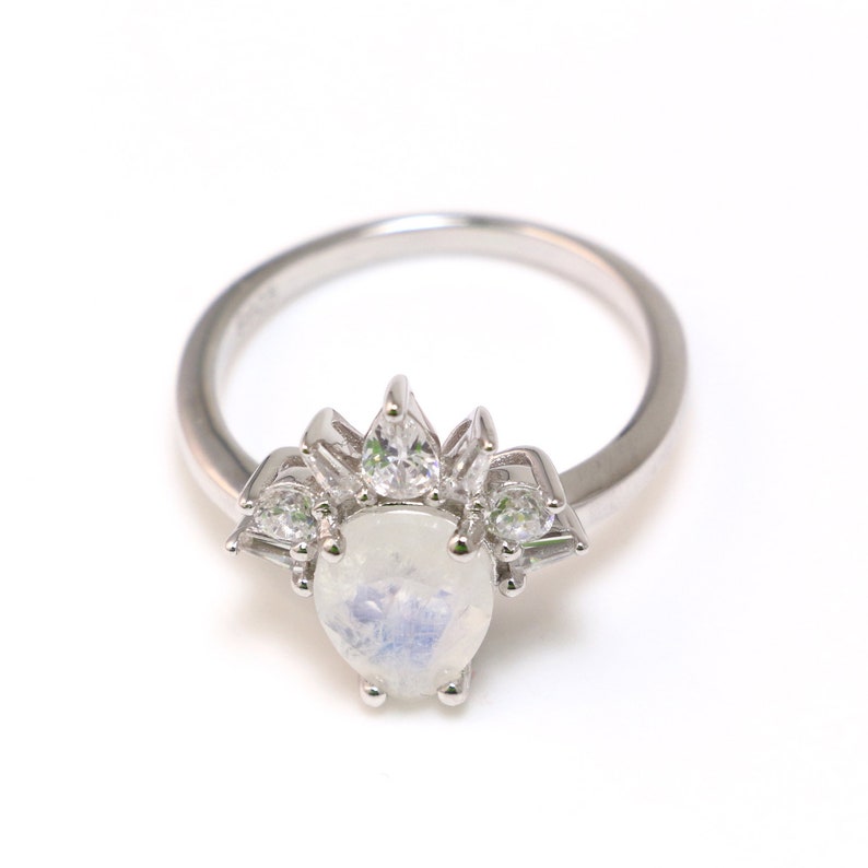 Puede incluir: Un anillo de plata con una piedra central de piedra lunar nacarada y piedras m&aacute;s peque&ntilde;as y claras alrededor.