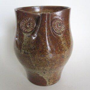 Puede incluir: Un jarrón de cerámica marrón con forma de búho hecho a mano con una superficie texturizada. El jarrón presenta detalles circulares en relieve que se asemejan a ojos. El jarrón con forma de búho es un artículo decorativo único.