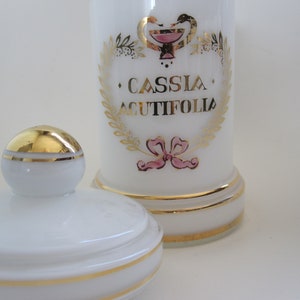 Puede incluir: Un tarro de boticario blanco con tapa y base doradas. El tarro presenta letras doradas y un emblema rosa y dorado, con las palabras "CASSIA ACUTIFOLIA" en un lugar destacado. El diseño incluye una corona dorada y un lazo rosa.