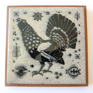 Op de afbeelding: Vierkante decoratieve tegel met een zwart-wit vogelontwerp, omlijst in hout. De vogel heeft een grote, waaierachtige staart en is omgeven door ster- en plantmotieven. De tegel heeft een textuur, gespikkelde achtergrond.