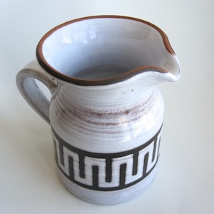 Puede incluir: Una jarra de cerámica con exterior blanco y marrón, con un patrón geométrico negro alrededor de la base. El borde y el asa son marrones. La jarra tiene un pico.