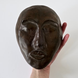 May include: A dark brown, sculpted face with closed eyes and a slightly parted mouth. The sculpture is held in a hand against a white background. The face has a smooth, matte finish.
