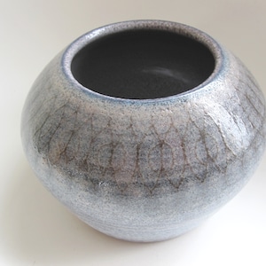 Może przedstawiać: Ręcznie wykonany ceramiczny wazon o zaokrąglonym kształcie i szerokim otworze. Wazon ma teksturowaną, nakrapianą powierzchnię w odcieniach szarości, niebieskiego i brązu. Obręcz jest ciemnoniebieska, a wnętrze czarne.