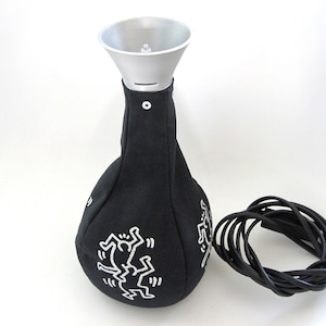 May include: A black fabric lamp with a silver metal funnel-shaped top. The black fabric body features white line art illustrations of figures in motion. A black electrical cord is coiled next to the lamp, which has a small white button fastener.