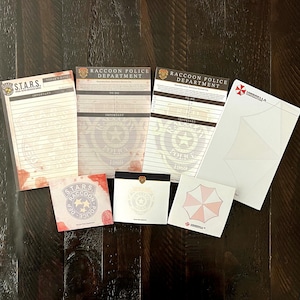 Puede incluir: Un conjunto de papelería que incluye tres blocs de notas rectangulares y tres blocs de notas adhesivas cuadrados. Los diseños presentan logotipos del Raccoon Police Department y S.T.A.R.S. con salpicaduras de sangre, y de Umbrella Corporation.