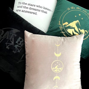 May include: Three decorative throw pillows with different designs. One pillow is black with a stylized bat design. Another pillow is white with black text that reads "To the stars who listen- and the dreams that are answered." The third pillow is beige with a gold design of a mountain range, stars, and crescent moons.