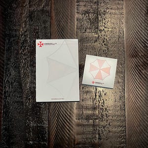 May include: A rectangular notepad and a square sticky note pad, both white with the red and white Umbrella Corporation logo. The large notepad features the text Umbrella Corporation and Our Business Is Life Itself. The small pad shows the logo.