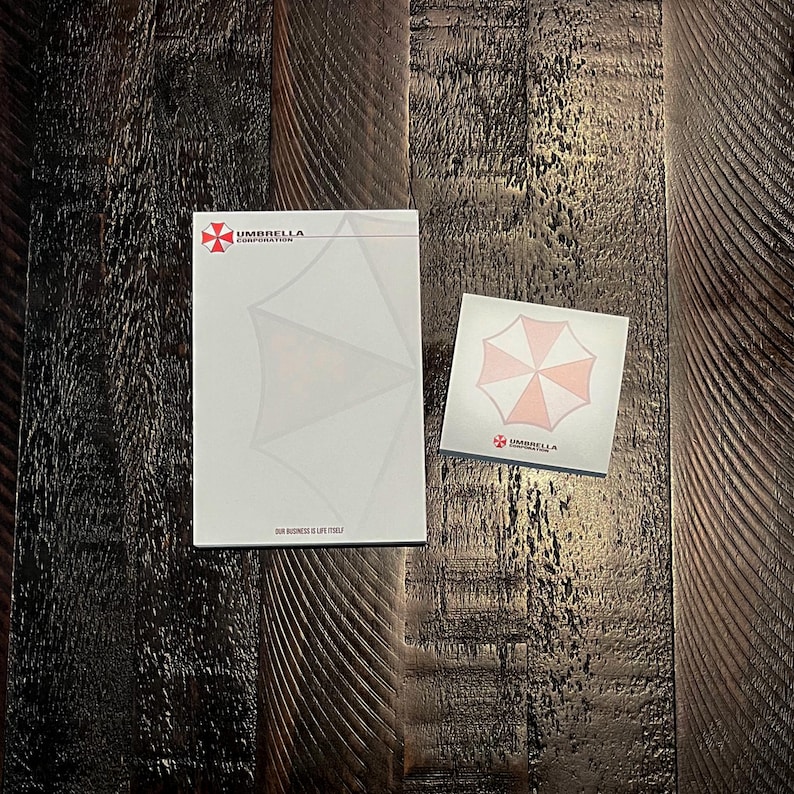 May include: A rectangular notepad and a square sticky note pad, both white with the red and white Umbrella Corporation logo. The large notepad features the text Umbrella Corporation and Our Business Is Life Itself. The small pad shows the logo.