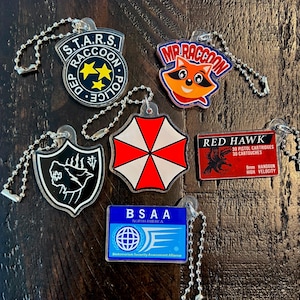 May include: A selection of acrylic keyrings with diverse designs. Includes a badge with "S.T.A.R.S. RACCOON POLICE", a raccoon face with "MR RACCOON", a red and white umbrella, and a "RED HAWK" cartridge design.