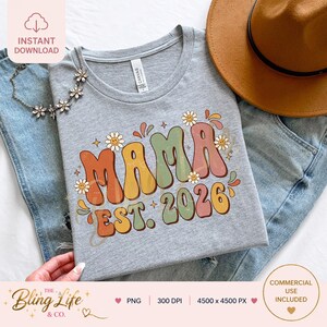以下が含まれることがあります： レトロなスタイルの「MAMA EST. 2026」という文字が入ったグレーのTシャツ。デザインは、カラフルで丸みを帯びた文字で「MAMA」という言葉と、デイジーのアクセントが特徴です。ジーンズと帽子と一緒に表示されています。