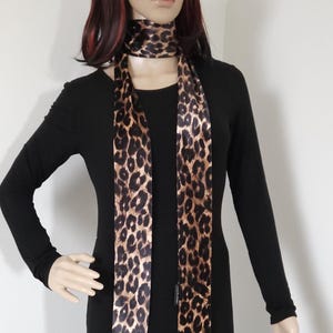 May include: A long, thin, brown and black leopard print satin scarf. The scarf is tied around the neck of a mannequin wearing a black dress.