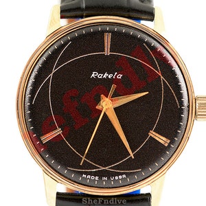 Może przedstawiać: Złoty zegarek na rękę z czarną tarczą i złotymi wskazówkami. Tarcza zegarka ma geometryczny wzór i napis "Raketa MADE IN USSR" nadrukowany na niej.