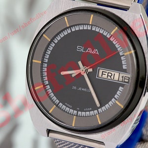 Puede incluir: Un reloj de pulsera Slava plateado con una esfera negra, detalles dorados y una ventana de fecha que muestra "FRI 18". El reloj tiene una correa de metal plateado y dice "26 Jewels Made in USSR".
