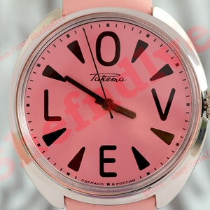 Puede incluir: Un reloj de pulsera rosa con caja plateada y correa de cuero rosa. La esfera del reloj tiene marcadores de hora en números romanos negros y una aguja de segundos negra. La esfera del reloj también tiene la palabra "Takema" impresa en negro. La esfera del reloj tiene el texto "Сделано в России" impreso en negro.