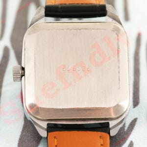 Puede incluir: Primer plano de un reloj de pulsera vintage. El reloj tiene una caja rectangular plateada con una corona plateada. La correa es de cuero negro con la parte inferior naranja. La parte trasera del reloj tiene el n&uacute;mero "606036" grabado en el metal.