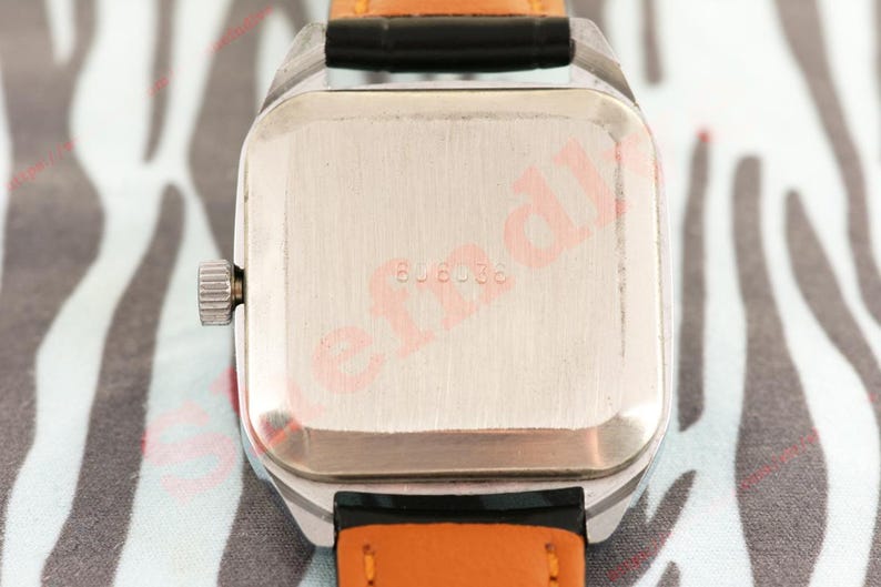 Puede incluir: Primer plano de un reloj de pulsera vintage. El reloj tiene una caja rectangular plateada con una corona plateada. La correa es de cuero negro con la parte inferior naranja. La parte trasera del reloj tiene el n&uacute;mero "606036" grabado en el metal.