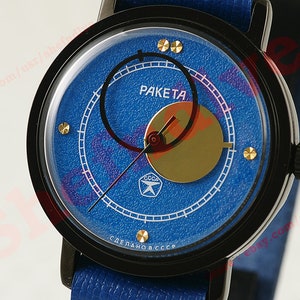 Puede incluir: Reloj Raketa azul con bisel negro y correa de cuero azul. La esfera del reloj presenta la palabra "Raketa" y un diseño dorado. El reloj tiene marcadores de hora dorados y una corona negra.