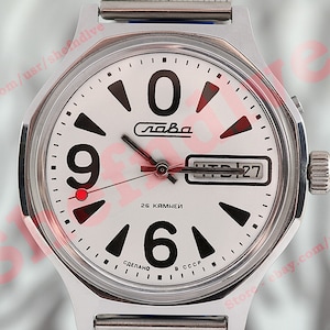 Puede incluir: Un reloj de pulsera plateado con una esfera blanca y números negros. El reloj tiene una ventana de fecha y un punto rojo en la posición de las 9 en punto. La esfera del reloj también dice "Слава" y "26 камней".